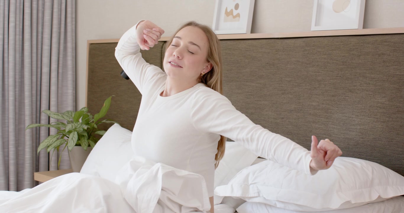 Female waking up, reaching for pink phone to start morning, sitting up on bed and stretching