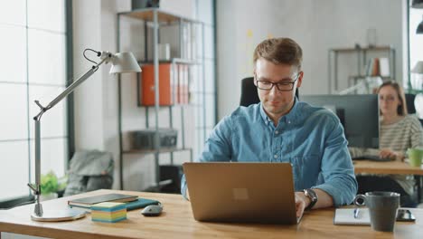 handsome young man in glasses and shirt is working on a laptop in a creative business agency. they work in loft office. diverse people working in the background. he's in good mood.