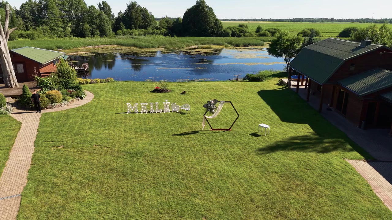 Aerial footage captures a tranquil lakeside venue with manicured green lawn, rustic wooden buildings, decorative 'MEILE' sign, blue sky, and bright natural sunlight. Ideal outdoor event setting.