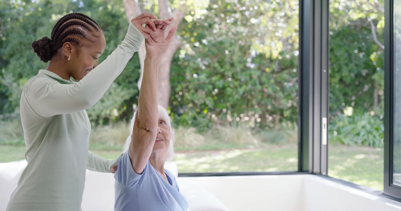 African American caregiver placing hands on shoulder guiding senior on chair by window doing rehab