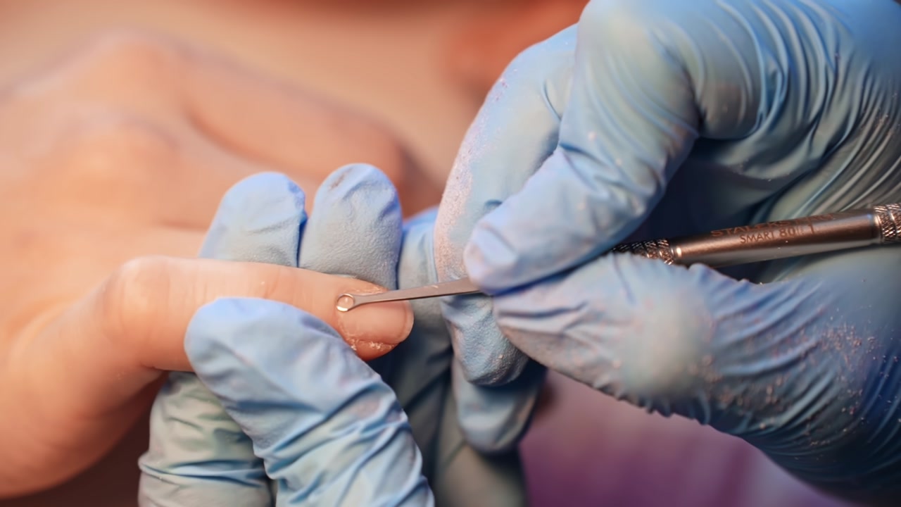 Close-up of a gloved nail technician using a rounded cuticle pusher to lift and clear the nail fold during careful professional manicure preparation.