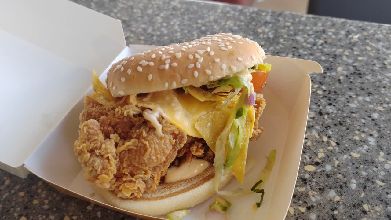 A Mexican-inspired burger with crispy chicken slice, cheese, and taco flavors served on a table inside a fast-food restaurant.