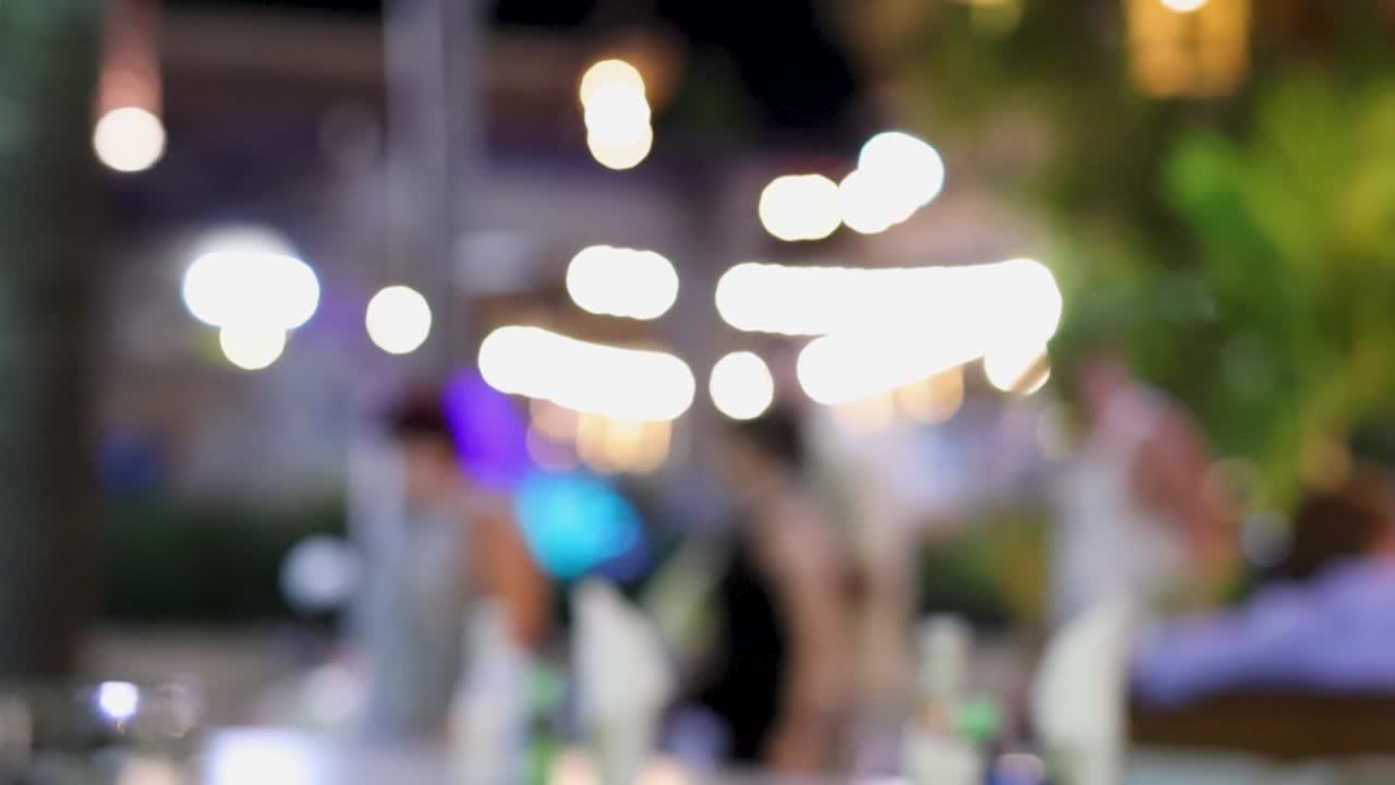 Out-of-focus scene with glowing lights and indistinct figures in an outdoor environment.
