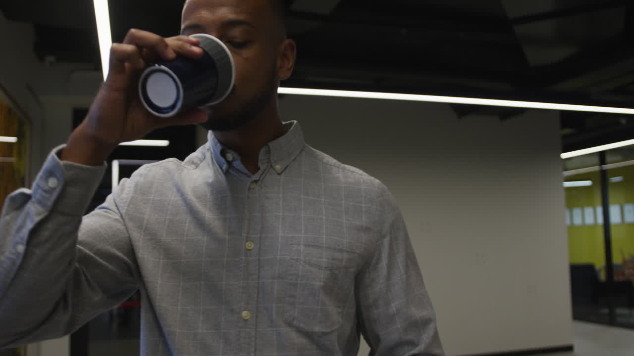 Empresário birracial caminhando e bebendo uma xícara de café em um escritório moderno