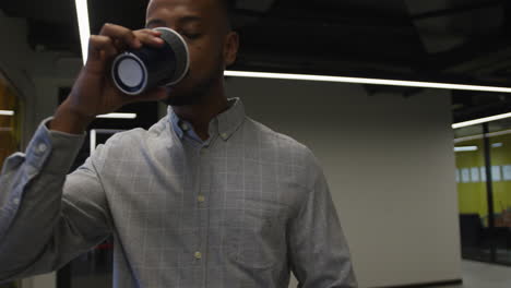 hombre de negocios biracial caminando y bebiendo una taza de café en una oficina moderna
