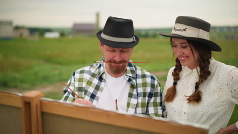 a scene of an artist focused on his work, holding a paintbrush in his mouth while wearing a checkered shirt. beside him, a woman in a white dress and hat stands, smiling as she gently touches his nose