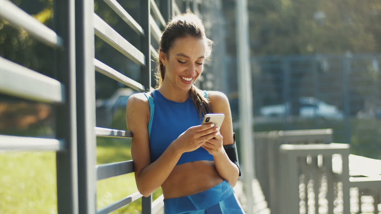 bonita deportista enviando un mensaje de texto en un smartphone y sonriendo en la cancha al aire libre en un día de verano