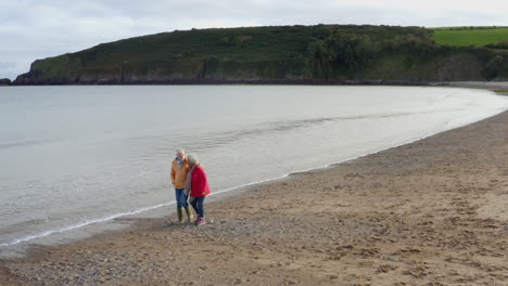 Drone-Shot-Of-Senior-Couple-Holding-Hands-As-They-Walk-Along-Shoreline-On-Winter-Beach-Vacation