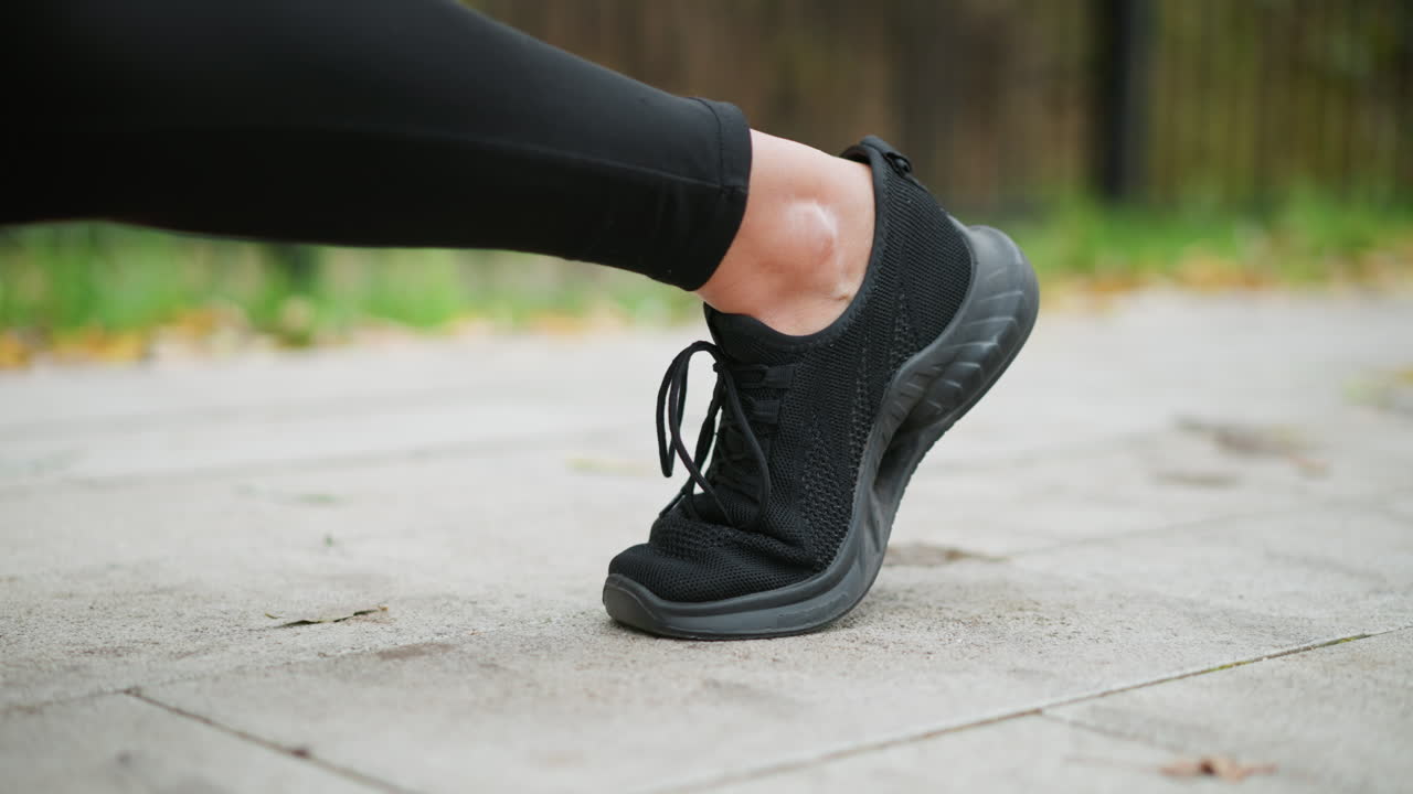Primo piano del piede e delle scarpe da ginnastica di un corridore con la gamba piegata, mentre si prepara per una corsa su un percorso asfaltato liscio, mettendo in risalto le calzature sportive con uno sfondo naturale sfocato.