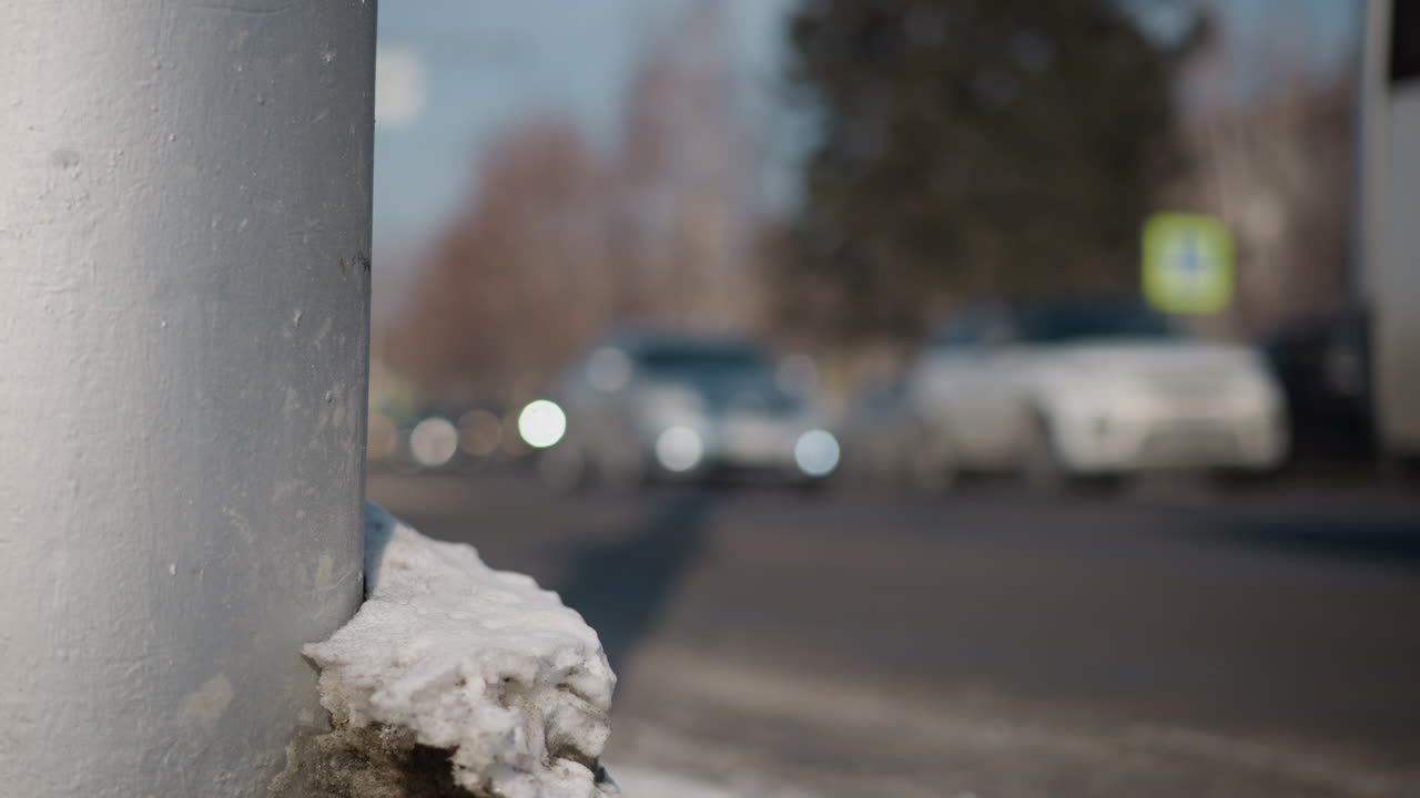 winter pole capped with snow near curb while blur cars and bus roll along city street with headlights glowing, soft focus bokeh behind, cold morning commute mood captured from roadside perspective