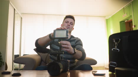 person sitting on sofa, holding in hand while examining camera gear setup on table. preparing for video shoot in home studio with camera, microphone, and lighting