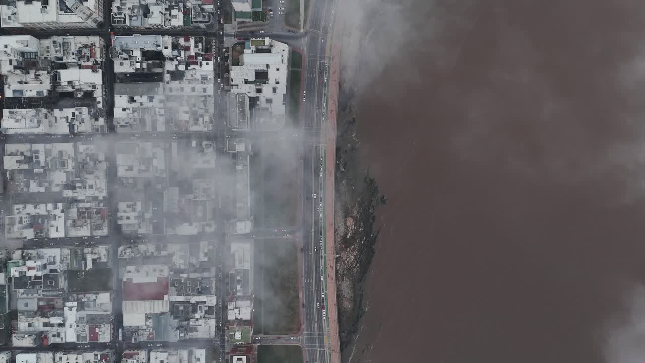 Aerial top-down view of Montevideo, featuring its iconic coastal boulevard (La Rambla), busy streets with cars in motion, and clouds drifting gracefully across the urban landscape