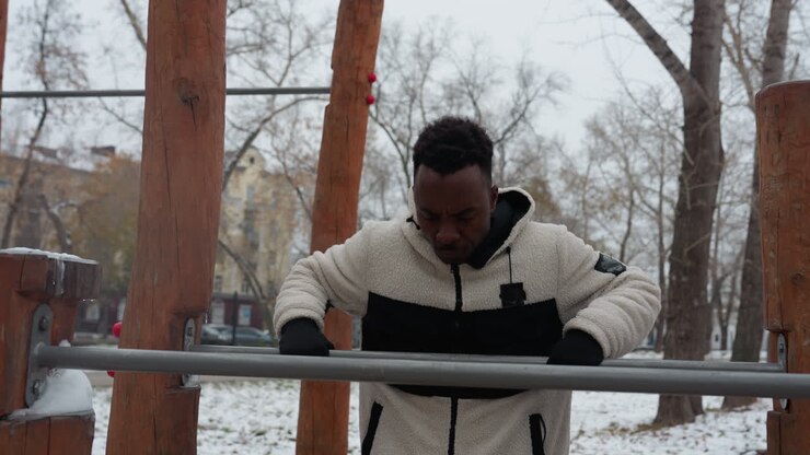Black boy in a winter jacket working out on outdoor gym equipment, focused and determined in a snowy urban park setting, surrounded by bare trees