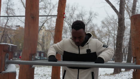 black boy in a winter jacket working out on outdoor gym equipment, focused and determined in a snowy urban park setting, surrounded by bare trees