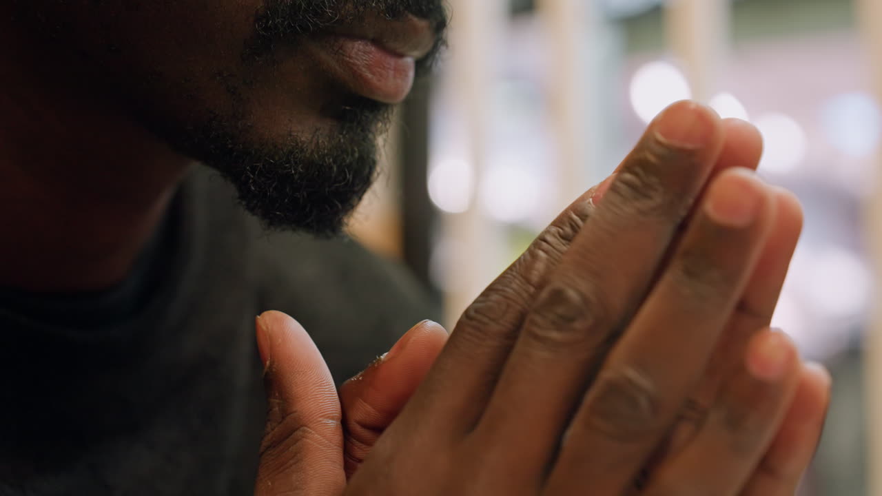 Black man praying hands close portrait, soft bokeh lights, folded fingers near lips, gentle beard, quiet indoor mood, intimate cinematic lighting, thoughtful meditation and gratitude, subtle motion