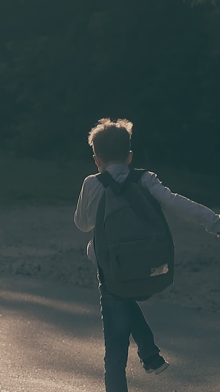 happy schoolboy runs skipping on sunny asphalt road with tree shadows and throws sheets slow motion backside view