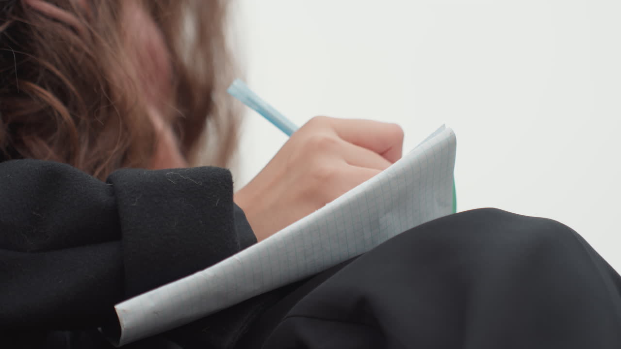 Studente che prende appunti in uno studio luminoso, primo piano delle mani che scrivono su un quaderno appoggiato sulle gambe, traccia di una penna color menta, manica di un cappotto nero, sfondo chiaro e delicato, calligrafia precisa, routine di studio organizzata e silenziosa
