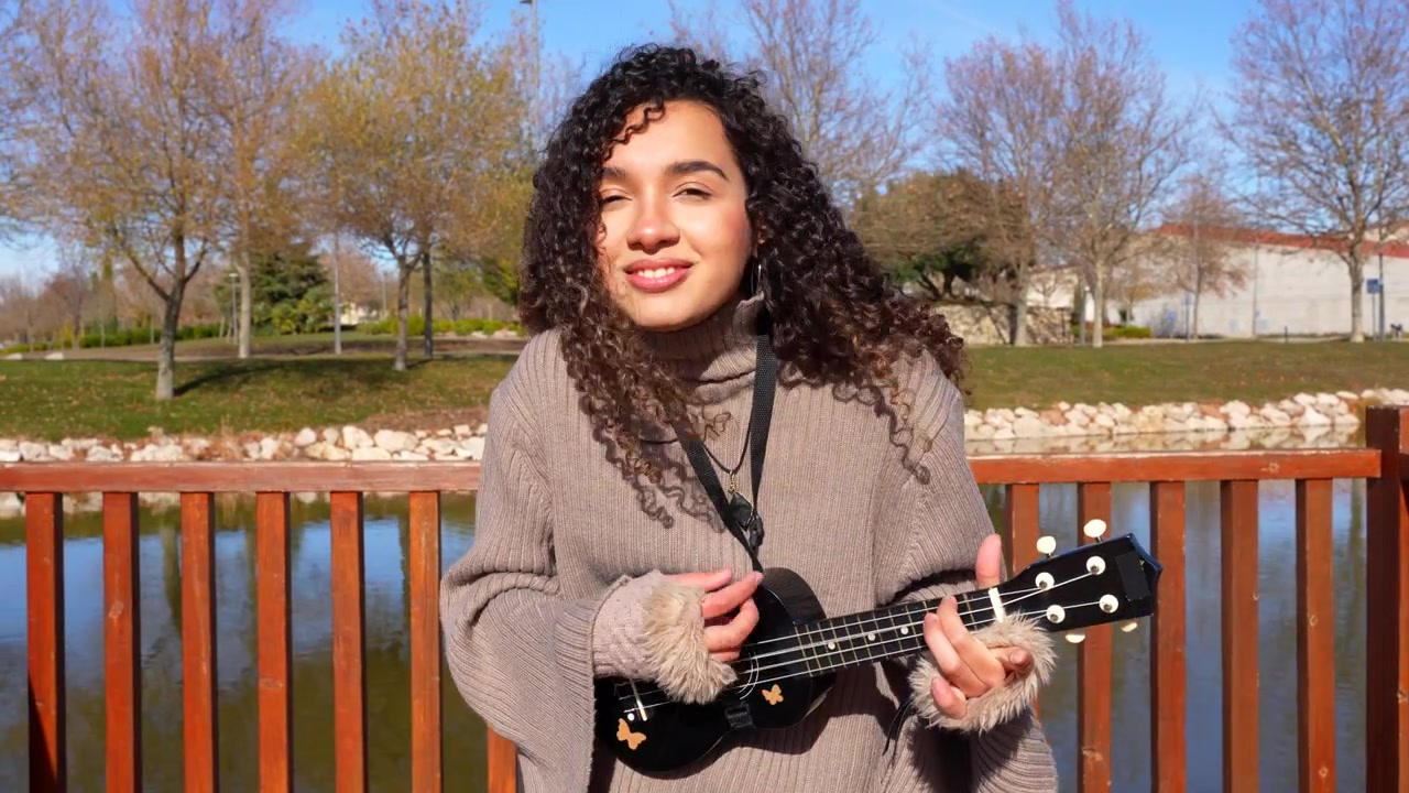 Cheerful young woman with curly hair playing the ukulele and singing happily in a park with a lake