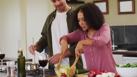 Happy-diverse-couple-preparing-a-meal-together-in-kitchen,-stirring-pan,-tossing-salad-and-laughing