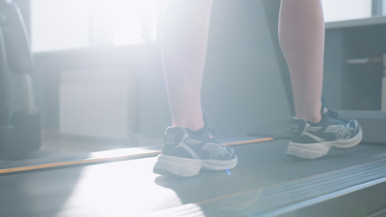lower angle of athlete walking on treadmill under sunlight illuminating her legs and sneakers in modern gym studio with exercise bikes and elliptical trainer blurred behind bright window light