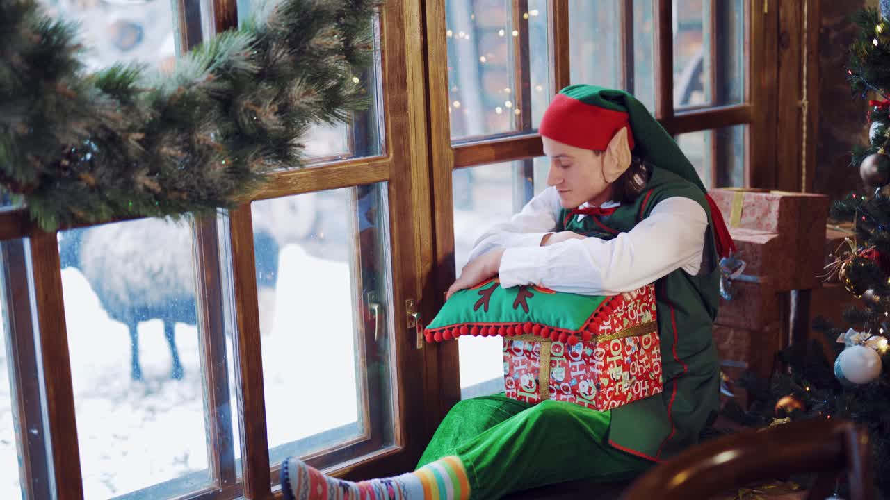Elf in a green suit sitting on the windowsill with a gift in his hand and a New Year's pillow and looking out the window at the deer waiting for a miracle on the background of the Christmas tree in the room during the day