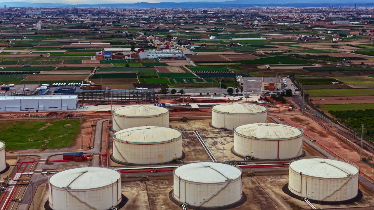 Tanques de armazenamento estão localizados em uma grande área industrial, perto de uma paisagem rural com campos e fazendas, durante o dia sob um céu claro.