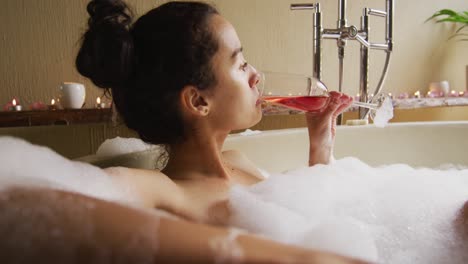 mujer biracial relajada acostada en un baño con espuma y mimándose con pétalos de rosa y vino
