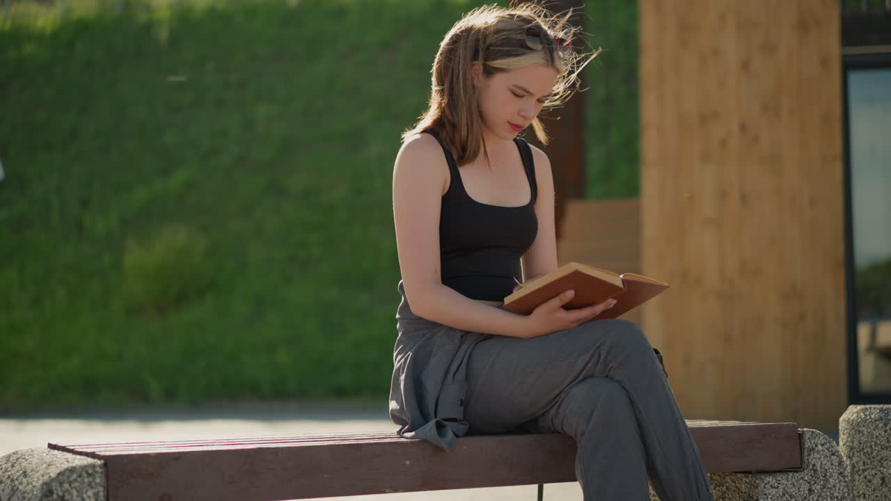 Lady in black singlet and grey joggers sits outdoors with crossed legs, reading a book, the wind blows her hair as she holds the book, and the sun reflects on her