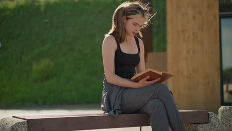 dama de soltera negra y corredores grises se sienta al aire libre con las piernas cruzadas, leyendo un libro, el viento sopla su cabello mientras sostiene el libro, y el sol se refleja en ella