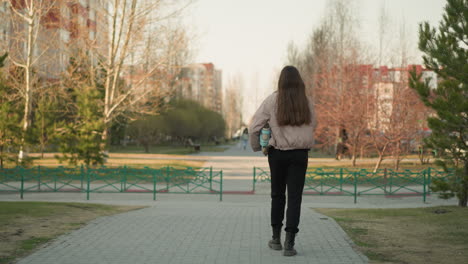 retrato de una chica con una chaqueta de melocotón, pantalones negros y botas negras, sosteniendo una patineta turquesa, caminando por un camino bordeado de árboles en un parque durante la primavera temprana