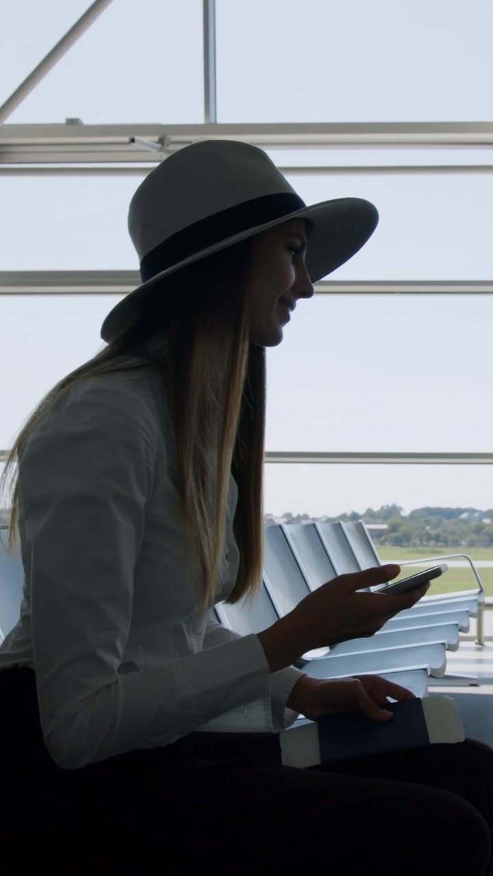A woman in a hat is sitting in an airport looking at her phone vertical video