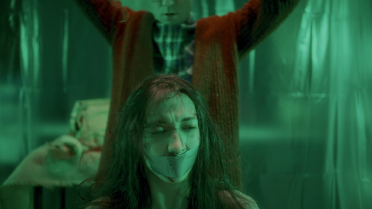 серийный убийца снимает мешок с головы напуганной жертвы с заклеенным ртом.