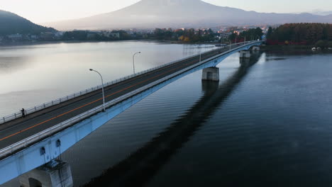 Vista-Aérea-Baja-Sobre-Un-Auto-En-El-Puente-Kawaguchiko,-Revelando-El-Monte-Fuji,-Brumoso,-Amanecer-De-Otoño-En-Japón