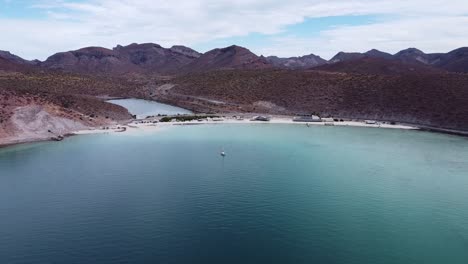 Aerial-wide-dolly-shot-of-the-beautiful-Playa-Pichilingue-near-Pichilingue-port-of-La-paz-in-Baja-California-Sur-Mexico-with-blue-sea-with-floating-ships-and-rocky-landscape-with-cloudy-sky