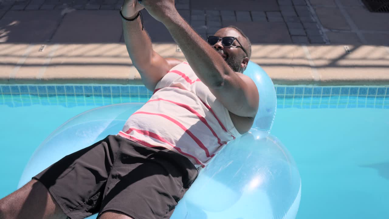 African American man in striped tank reclining on pool float glancing at phone framing selfie