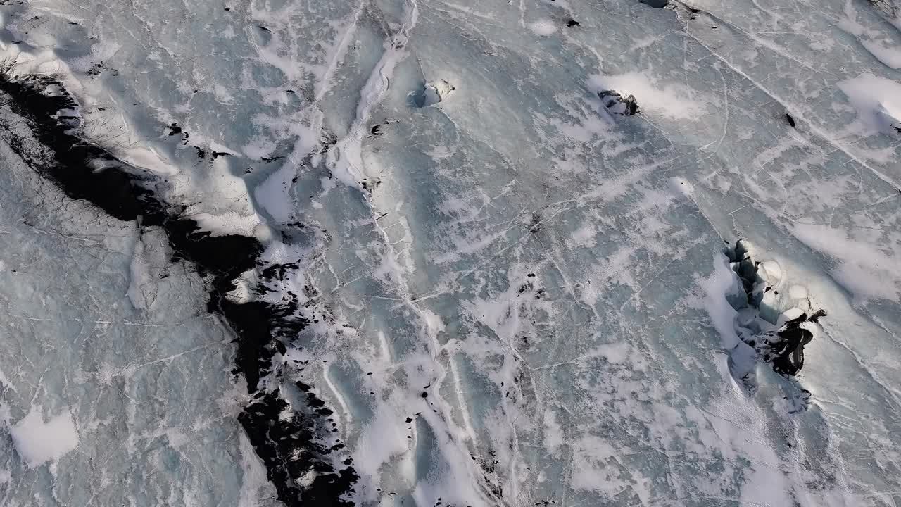 Top-down aerial view of the frozen, textured surface of a glacier in Iceland, showcasing intricate ice patterns, dark crevasses, and frosty landscapes under cold light.