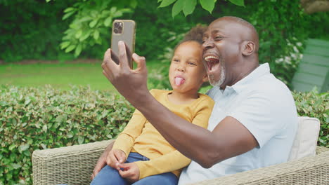 Grandfather-Pulling-Faces-With-Granddaughter-Taking-Selfie-On-Mobile-Phone-In-Garden-At-Home