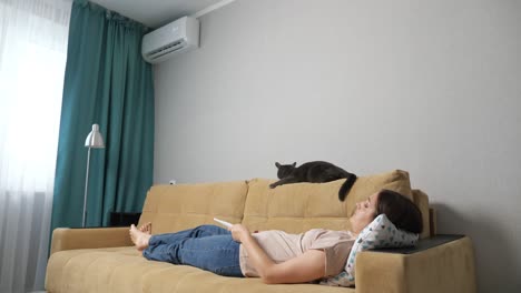 woman turns on the air conditioner by the remote control, lying on the couch with a cat