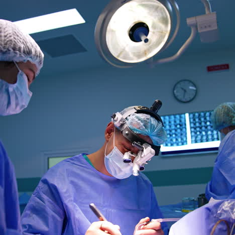 caucasian surgeon in uniform and device glasses sits near the patient. doctor uses metal tool energetically. assisting staff around the doctor.
