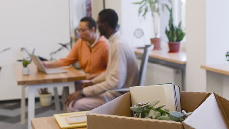 close up view of a cardboard house with personal belongings, businesswoman talks to a young employee while they look at the computer sitting at the table in the background