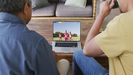 Video-Von-Vater-Und-Sohn,-Die-Auf-Der-Couch-Sitzen-Und-Sich-Ein-Fußballspiel-Auf-Dem-Laptop-Ansehen