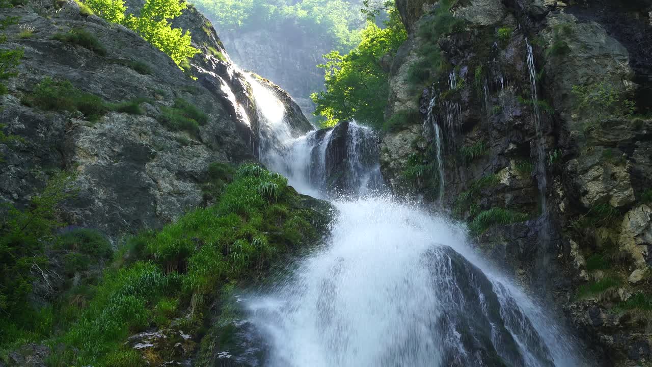 Arnavut Alpleri'nin yüksek dağlarında gün doğumu, kayalık yamaçlı ve yeşil ağaçlı şelale manzarası