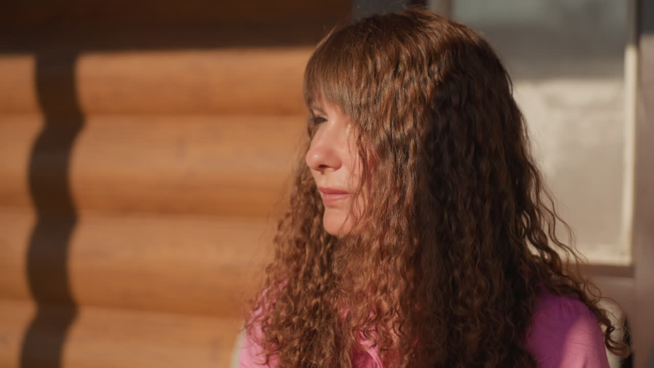 Reflective Woman With Curly Hair Eating Outdoors, Caucasian Woman With Pink Top Relaxing On Cabin Porch, Serene Woman With Curly Hair At Rustic Cabin Enjoying Quiet Breakfast In Sunlight