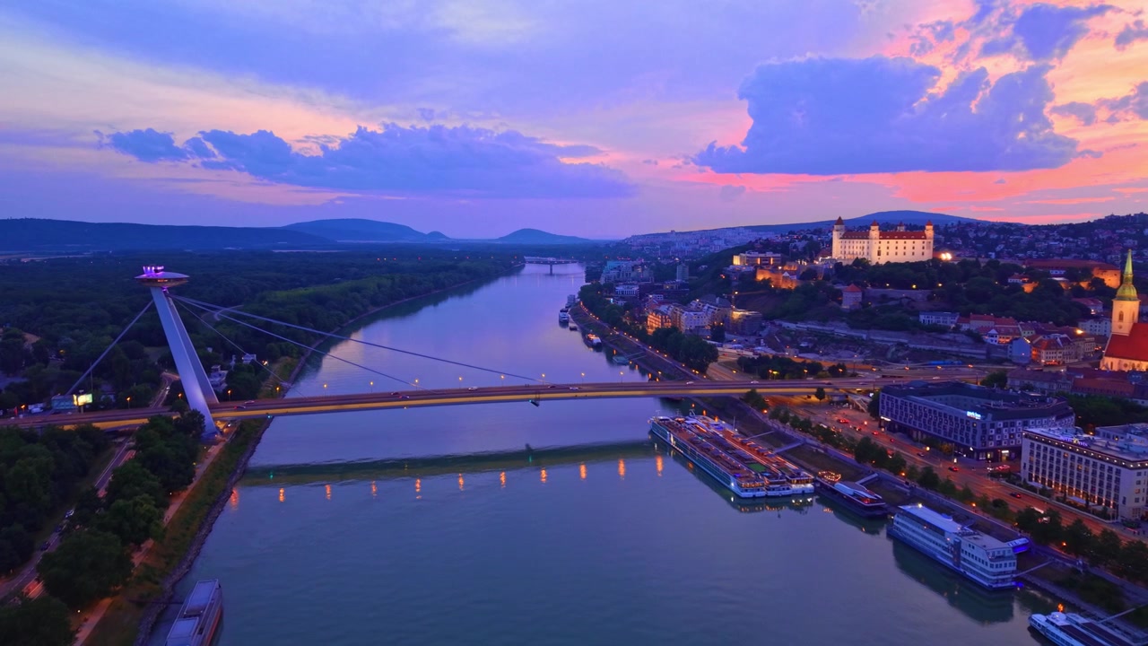 Vue de Bratislava et du Danube. La ville est présentée au coucher du soleil, avec le fleuve, ses ponts et les bâtiments qui bordent ses deux rives.