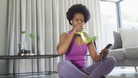 feliz afroamericano usando ropa deportiva, usando el teléfono inteligente, bebiendo agua