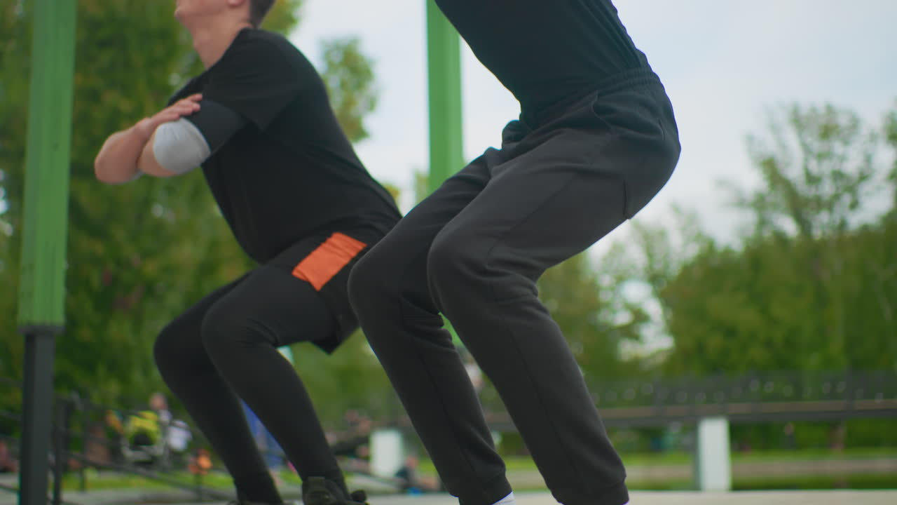 Duo che si allena con squat in un parco all'aperto, coach che guida l'atleta attraverso squat profondi controllati, alberi verdi sullo sfondo, ringhiera di metallo e panchina urbana, abbigliamento sportivo nero casual con dettagli arancioni sulle tasche