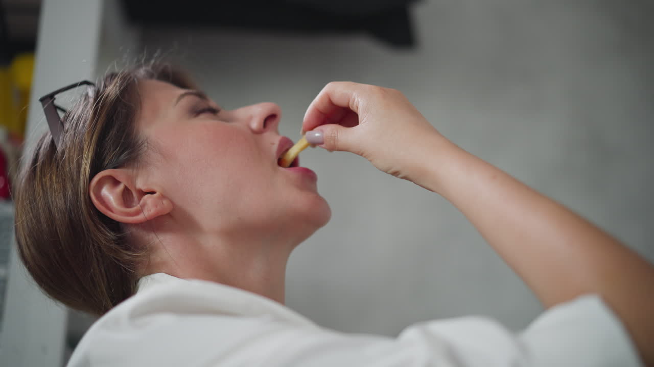 Playful woman wearing white top raises head slightly while eating chips dipped in sauce indoors with blurred background visible showing relaxed casual moment during break in cozy home