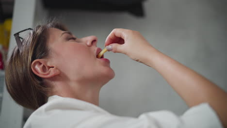 playful woman wearing white top raises head slightly while eating chips dipped in sauce indoors with blurred background visible showing relaxed casual moment during break in cozy home