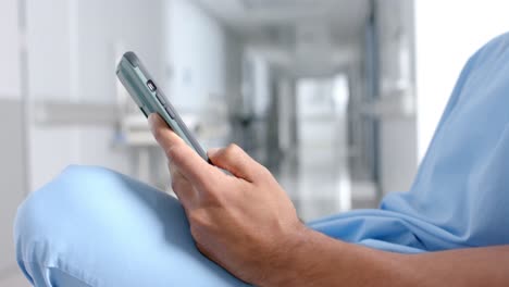 sección media de un médico masculino biracial usando un teléfono inteligente en el pasillo, en cámara lenta