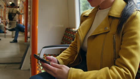 Close-Up-Of-Young-Woman-Sitting-On-Underground-Train-On-Journey-To-Work-Checking-Text-Alert-On-Mobile-Phone